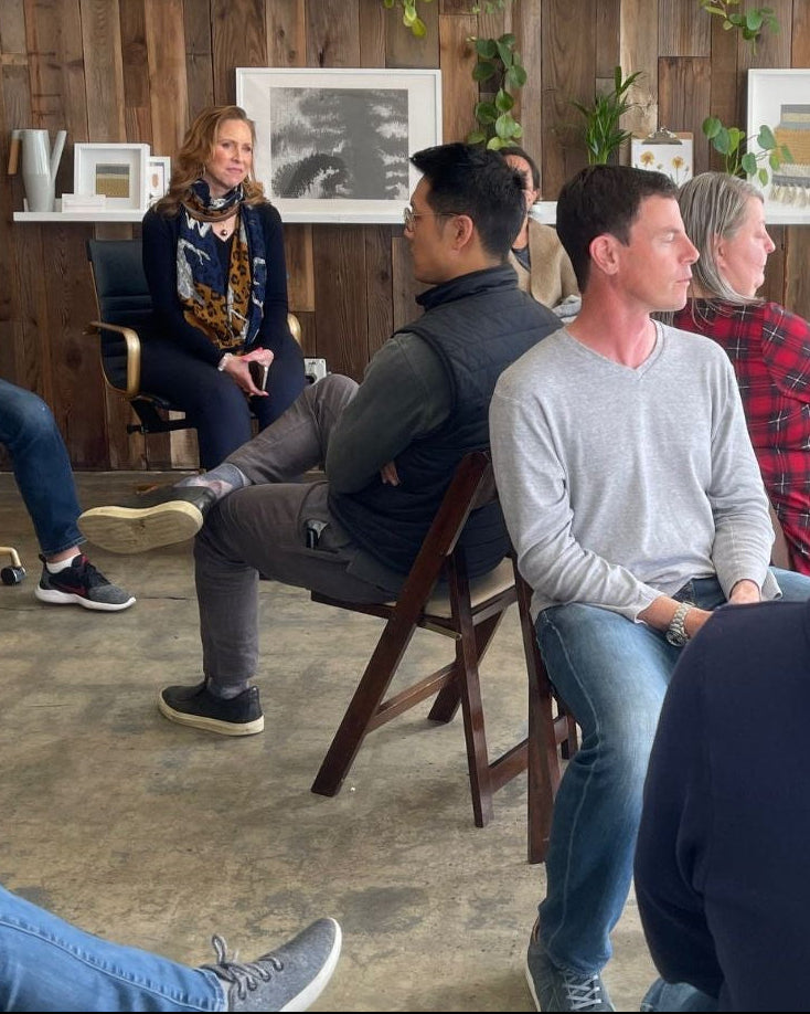 Group of people sitting in a circle in a room with wooden walls and plants.